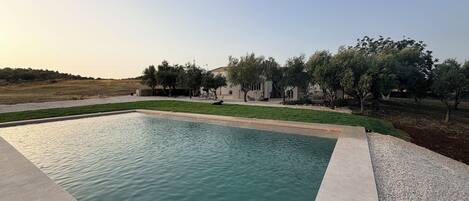 A swimming pool with a surrounding patio area, a grassy lawn, and a building with trees in the background.