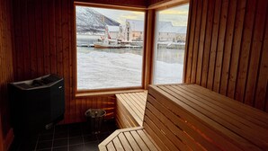 Una sala de sauna con paredes y bancos de madera, una estufa de sauna y un ventanal con vistas a un paisaje nevado y barcos.