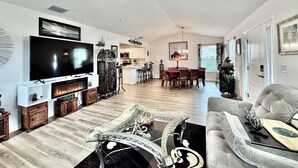 A spacious living room with a fireplace, a large flat-screen TV, a ceiling fan, and a dining area with a table and chairs.