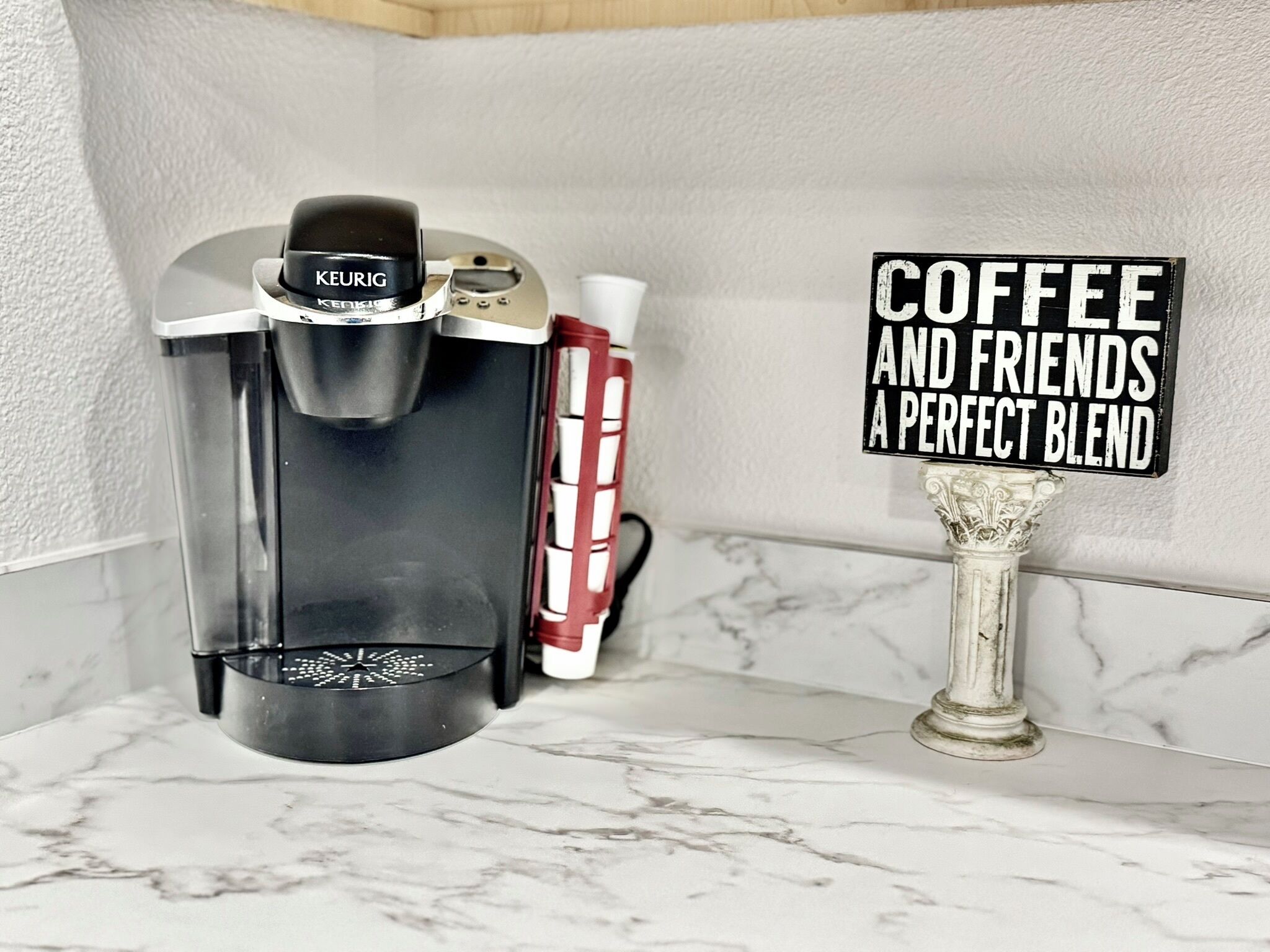 Una cafetera, un termo vermell i blanc, i un cartell que diu "COFFEE AND FRIENDS A PERFECT BLEND" sobre una superfície de marbre.