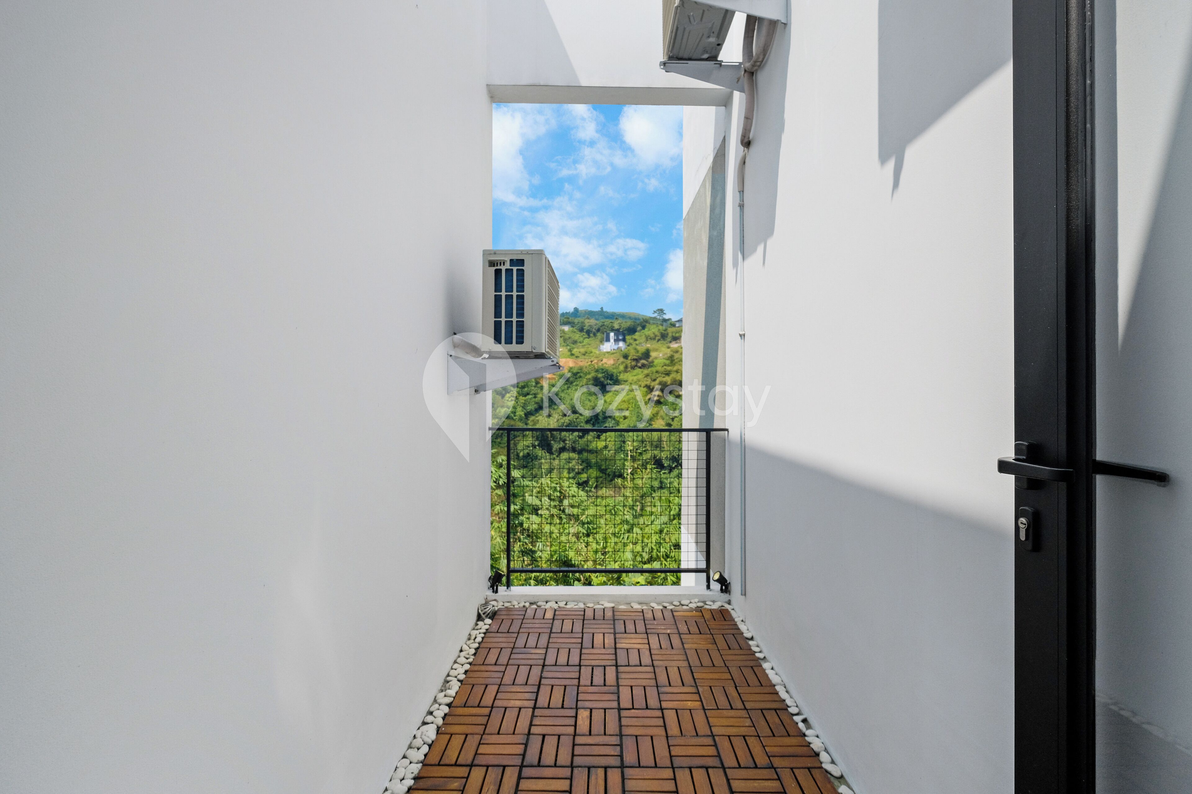 A narrow corridor with a wooden floor, a black door, and a window with a view of greenery and a building.