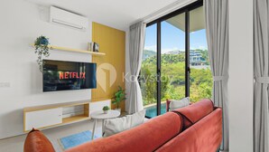 A modern living room with a flat-screen TV, a red sofa, a small table with a plant, and a view of greenery and buildings through large windows.