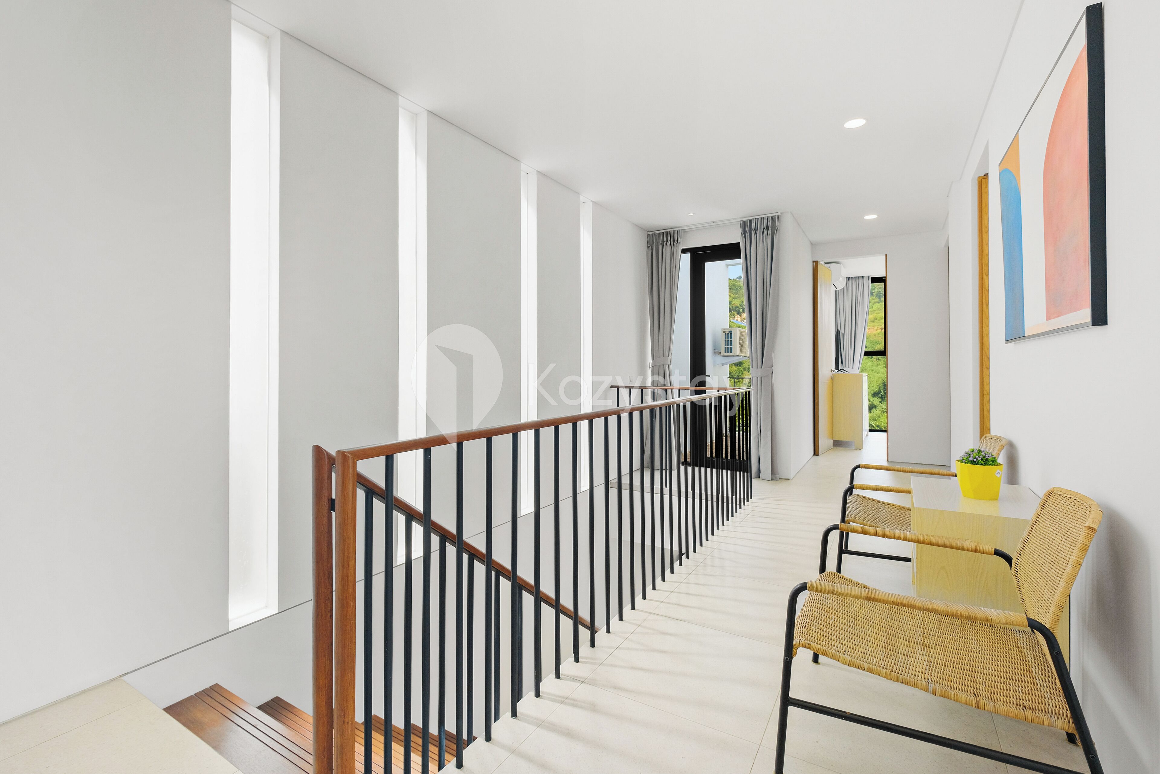 A modern interior with a staircase, a dining area with wicker chairs, and a view of greenery outside.