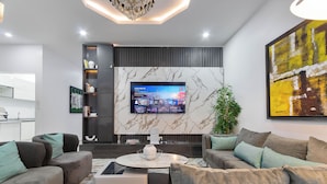 Een moderne woonkamer met een flatscreen televisie bevestigd aan een marmeren muur, grijze banken met turquoise kussens, een salontafel en een potplant.