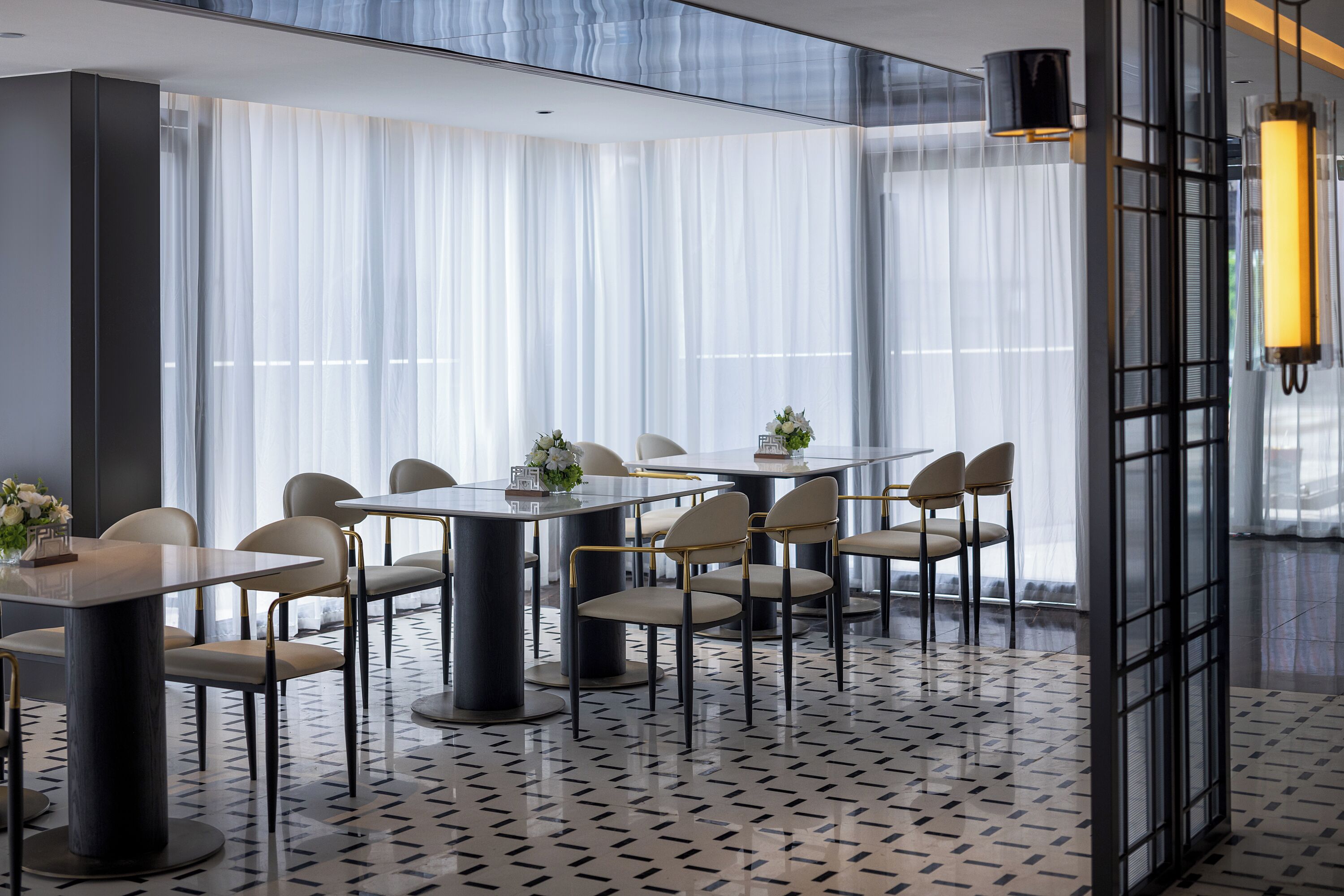 A modern hotel room with a large window, a dining area with round tables and chairs, and a patterned floor.