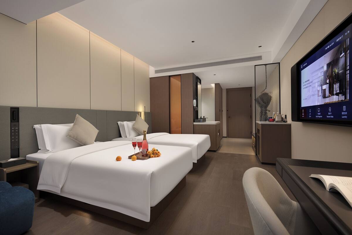 Habitación de hotel moderna con dos camas, un escritorio, televisión y baño visible a través de una puerta abierta.