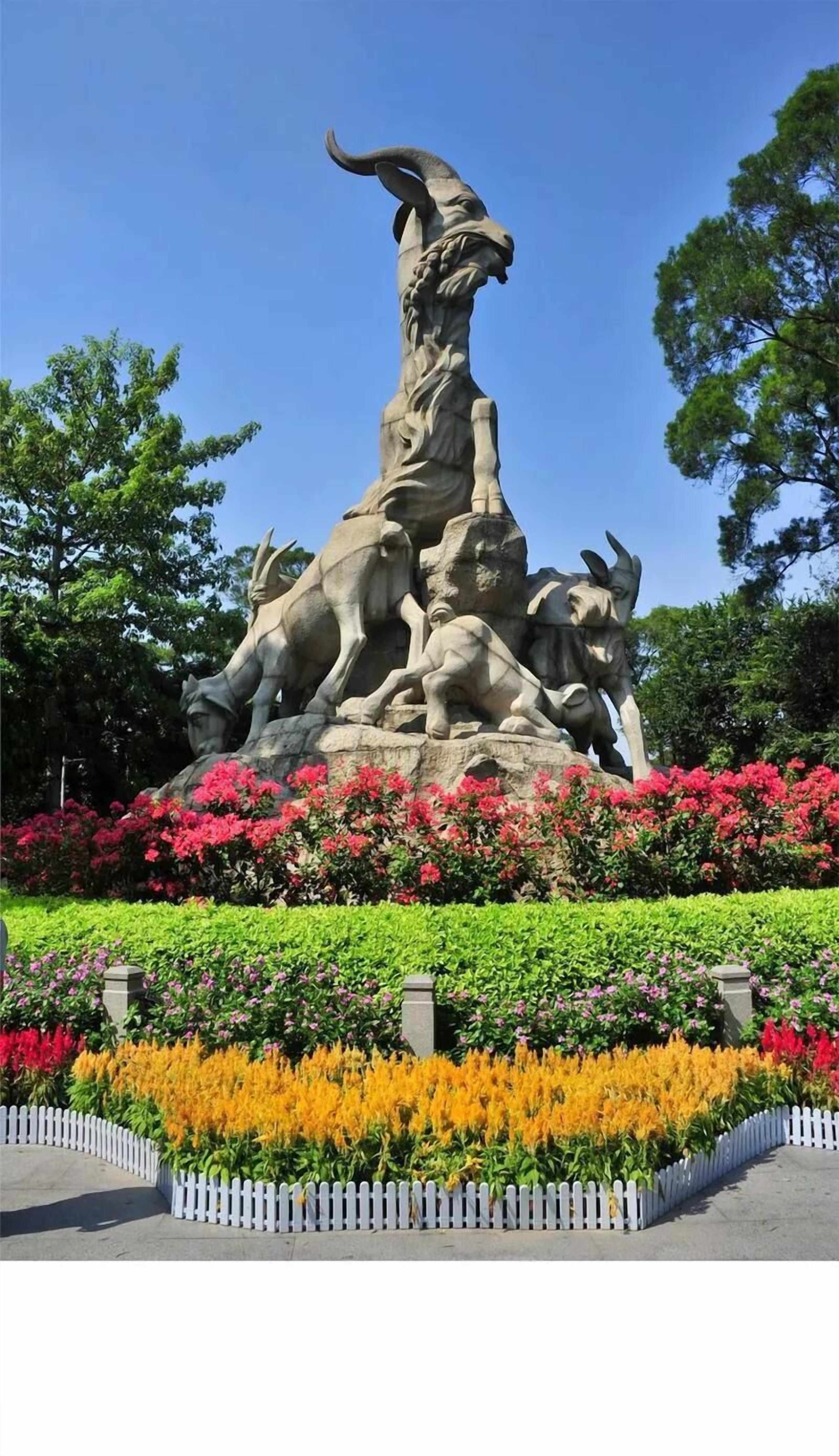 A large outdoor sculpture with a dynamic figure, surrounded by a variety of colorful flowers and a white picket fence.
