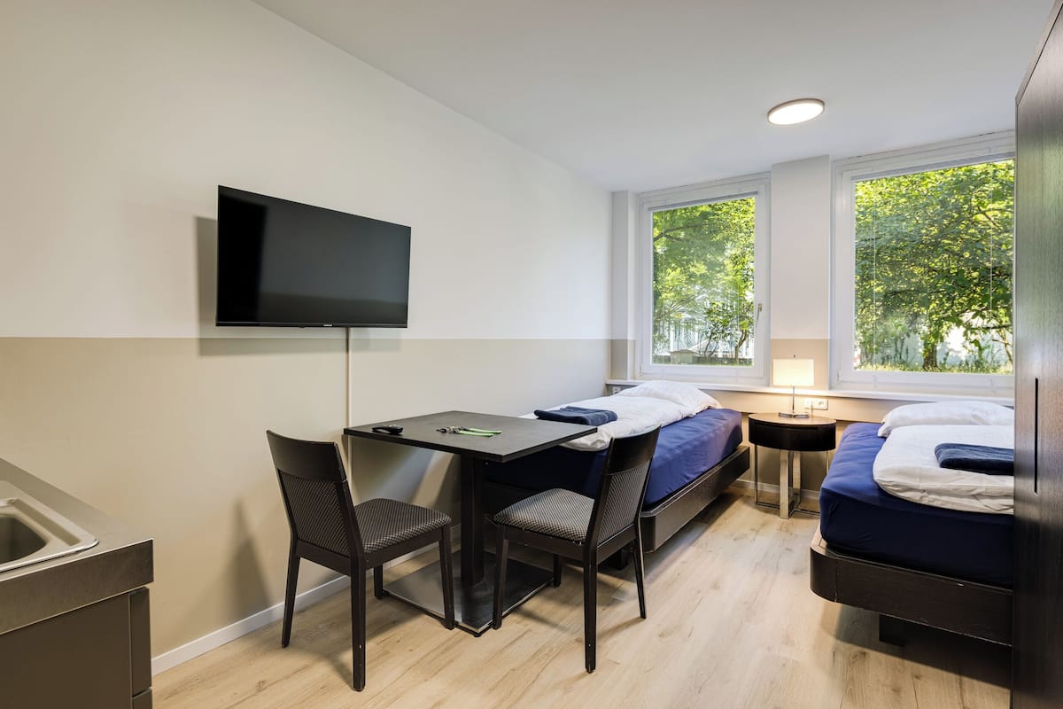 A compact room with a bed, a desk with two chairs, a television, and a window with a view of trees.
