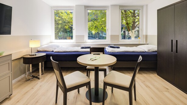 Una habitación moderna y funcional con cama, una mesita pequeña con dos sillas, un escritorio con lámpara y un armario.