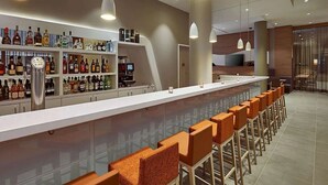 Een moderne bar met een lange toonbank, meerdere rekken vol drankflessen en verschillende oranje barkrukken.