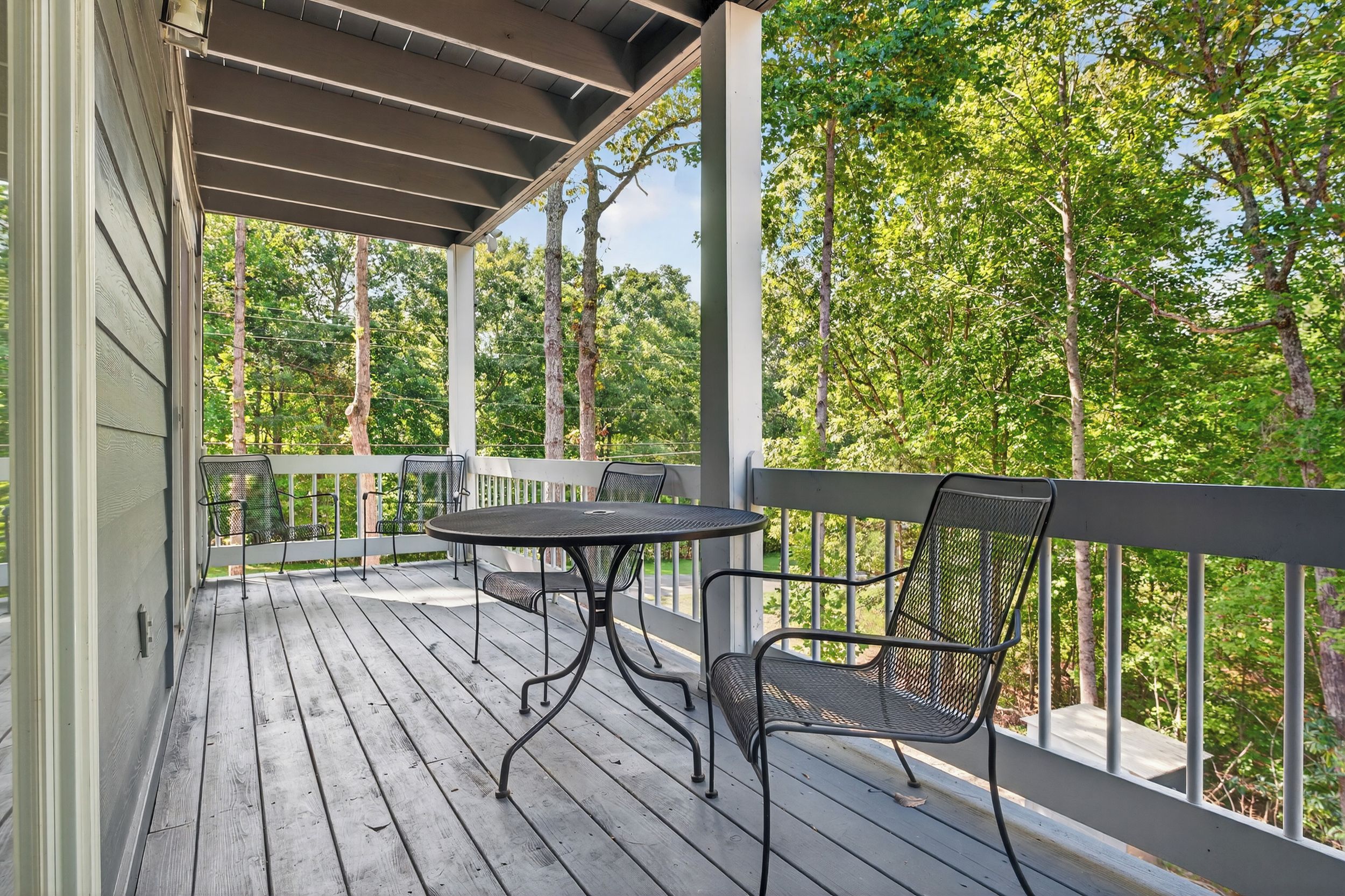A wooden deck with a table and chairs, surrounded by a railing, overlooking a forested area.