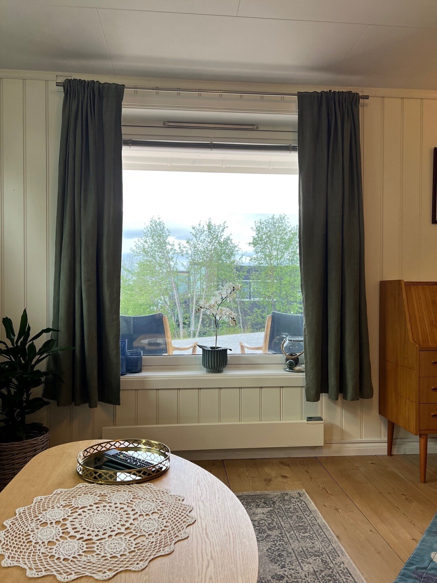 A room with a large window, a wooden table with a lace doily, a small potted plant, and a wooden cabinet.
