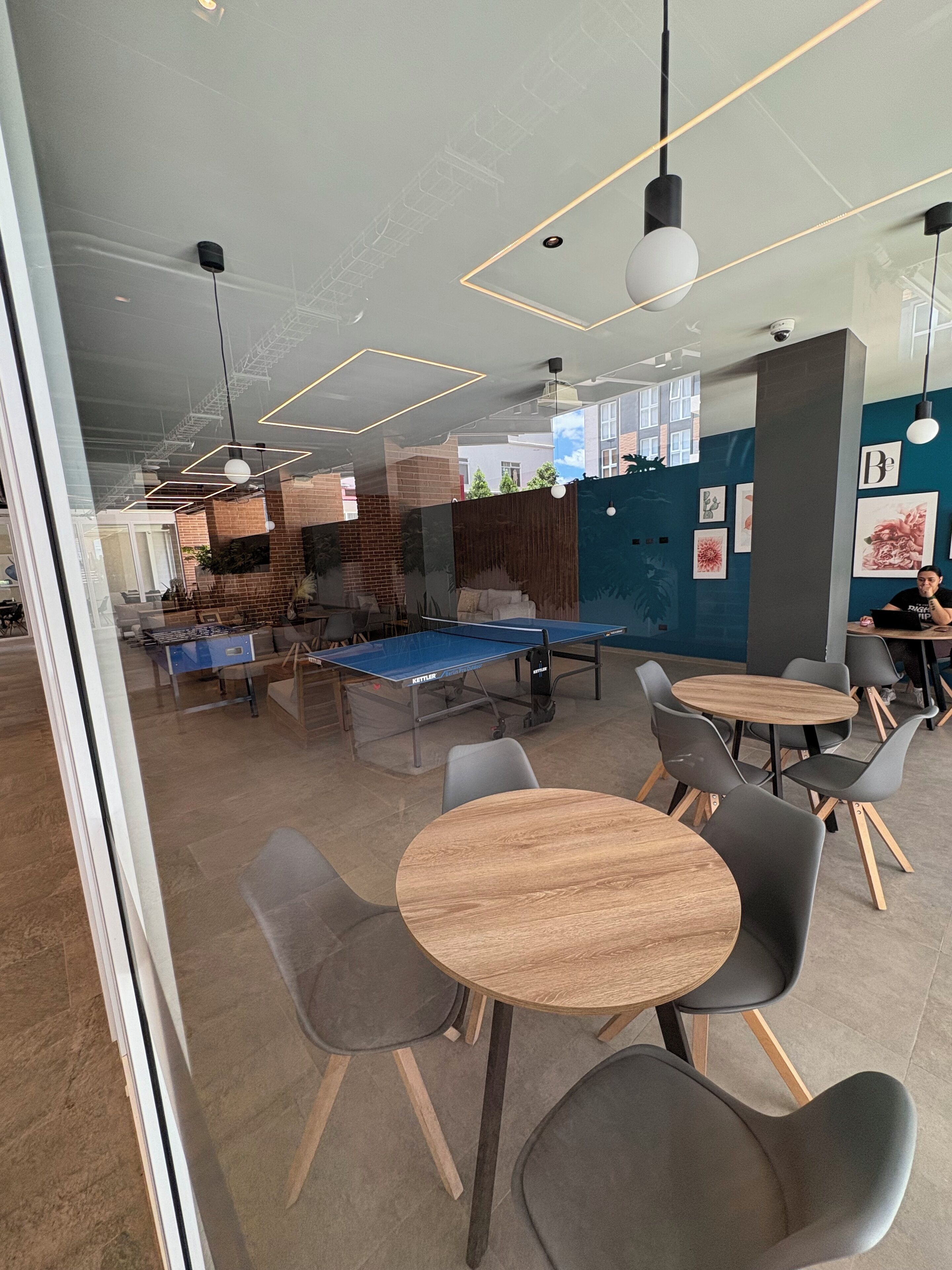 A modern indoor space with ping pong tables, round wooden tables, and grey chairs.