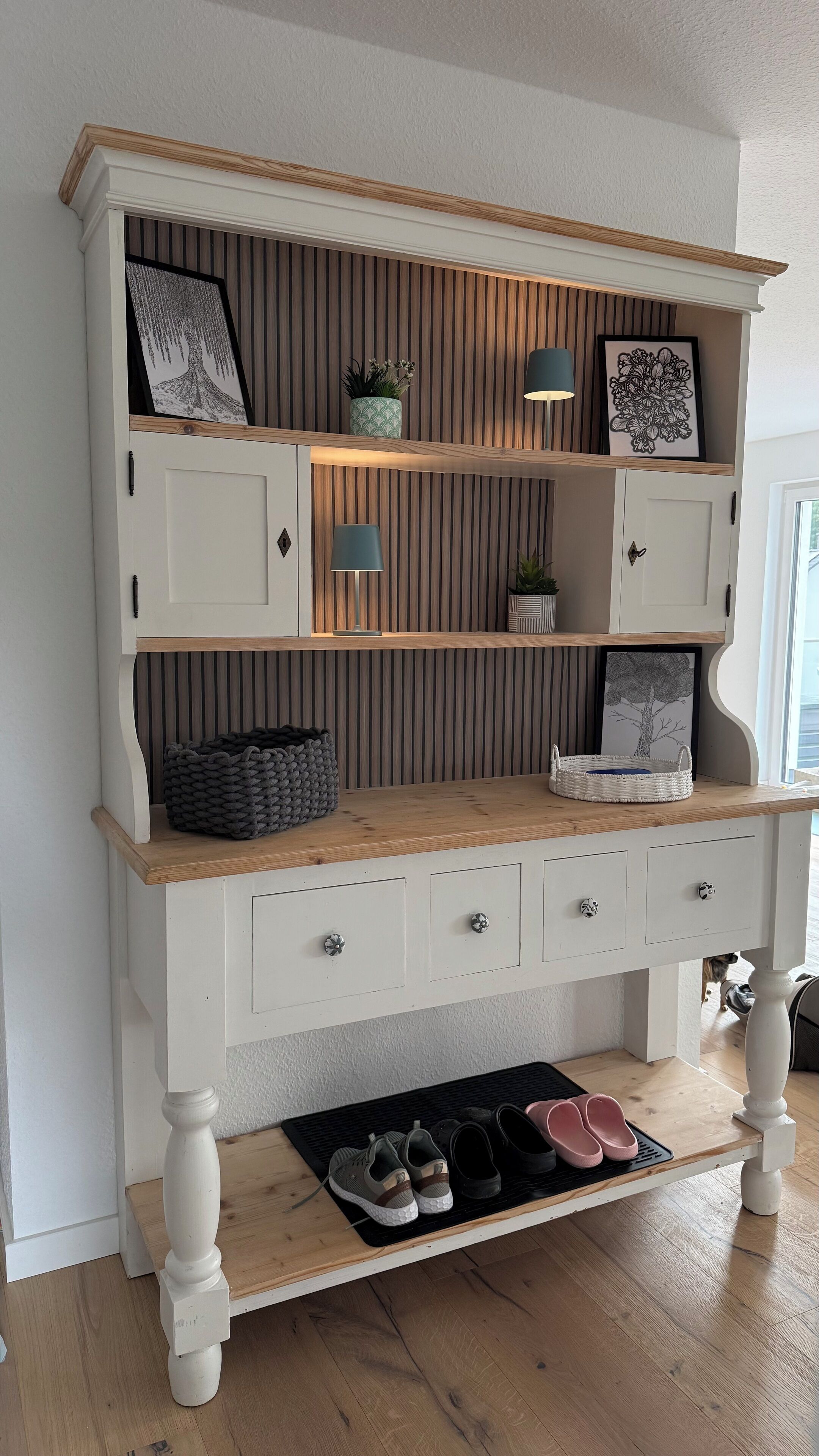 A white wooden bench with shelves and drawers, featuring framed artwork, potted plants, and a tray with shoes.