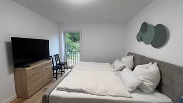 Camera da letto con un letto, una TV su una cassettiera in legno, una sedia e un balcone con una porta.