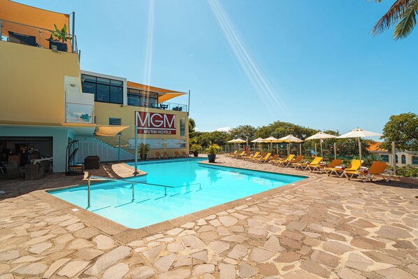 A swimming pool area with lounge chairs and umbrellas, a building with a 'MGM' sign, and a stone-tiled deck. - Blissful Getaway! Mountain View, Swimming Pool, Spa & Wellness, Free Parking! (Funchal)