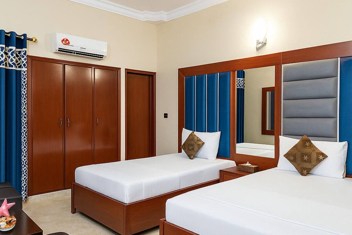 A hotel room with two beds, a wardrobe, an air conditioning unit, a desk with a chair, and a ceiling fan.