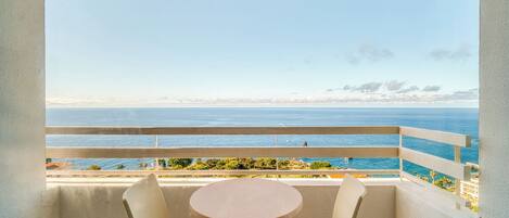 A balcony with a table and chairs, overlooking a coastal view with a clear sky.