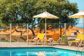 A poolside area with lounge chairs and umbrellas, a clear pool, and a view of a building with orange-tiled roofs. - Ultimate Beach Escape in Madeira! Outdoor Pool, Free Parking, Mountain View! (Funchal)