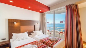 A hotel room with two beds, a wooden headboard, a large window with a view of the sea, and a balcony with chairs.