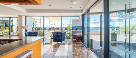 A modern hotel lobby with a reception desk, seating area, and large windows offering a view of the ocean.