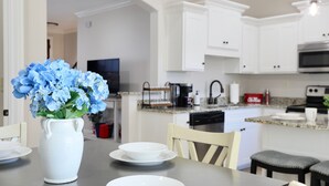 A modern kitchen with white cabinets, a granite countertop, and a dining area with a vase of blue flowers. - "Swayze Stays" - Charming Oxford Condo! (Oxford)