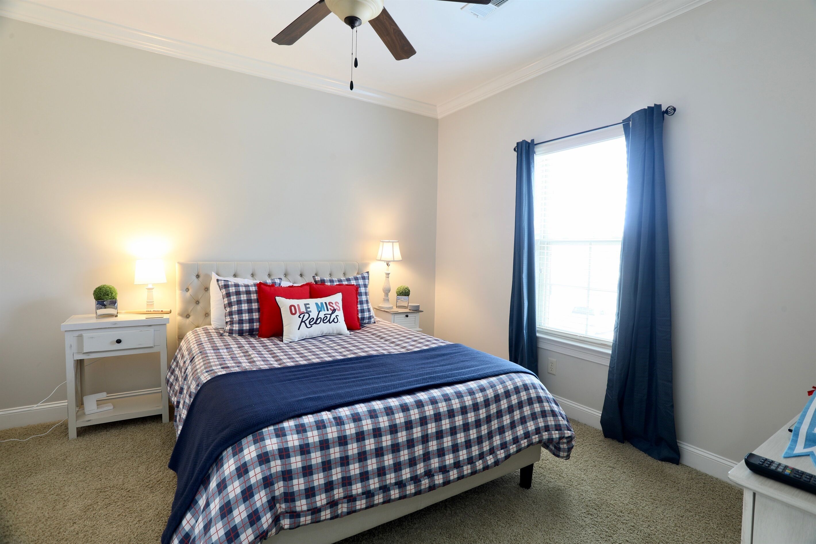 A bedroom with a bed, nightstands, a ceiling fan, a window with curtains, and a lamp.