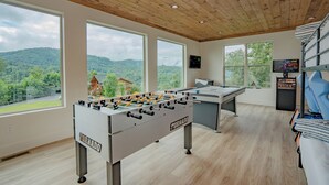 A modern room with a foosball table, large windows, and a view of the outdoors. - 🏔️ Apres All Day 🌙 Rooftop Mountain Views, Putting Green, Hot Tub & Arcade (Gatlinburg)