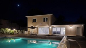Una piscina con vista despejada del área de la piscina, que incluye una mesa y sillas bajo una pérgola, y una casa con ventanas iluminadas y un balcón al fondo.