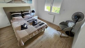 Un dormitorio con cama, una silla, un ventilador de pie, una mesita de noche y una ventana con persianas.