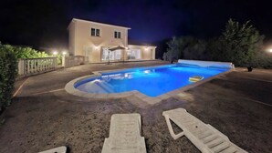 Una piscina con sillones de descanso, una casa y una zona de patio bien iluminada por la noche.