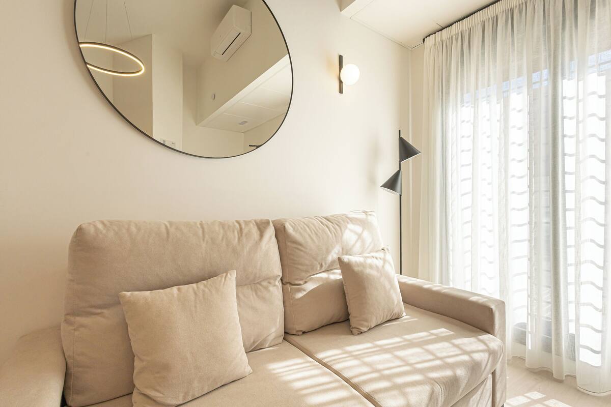 A neutral-toned sofa with cushions, a round mirror on the wall, a wall-mounted light, and sheer curtains allowing natural light.