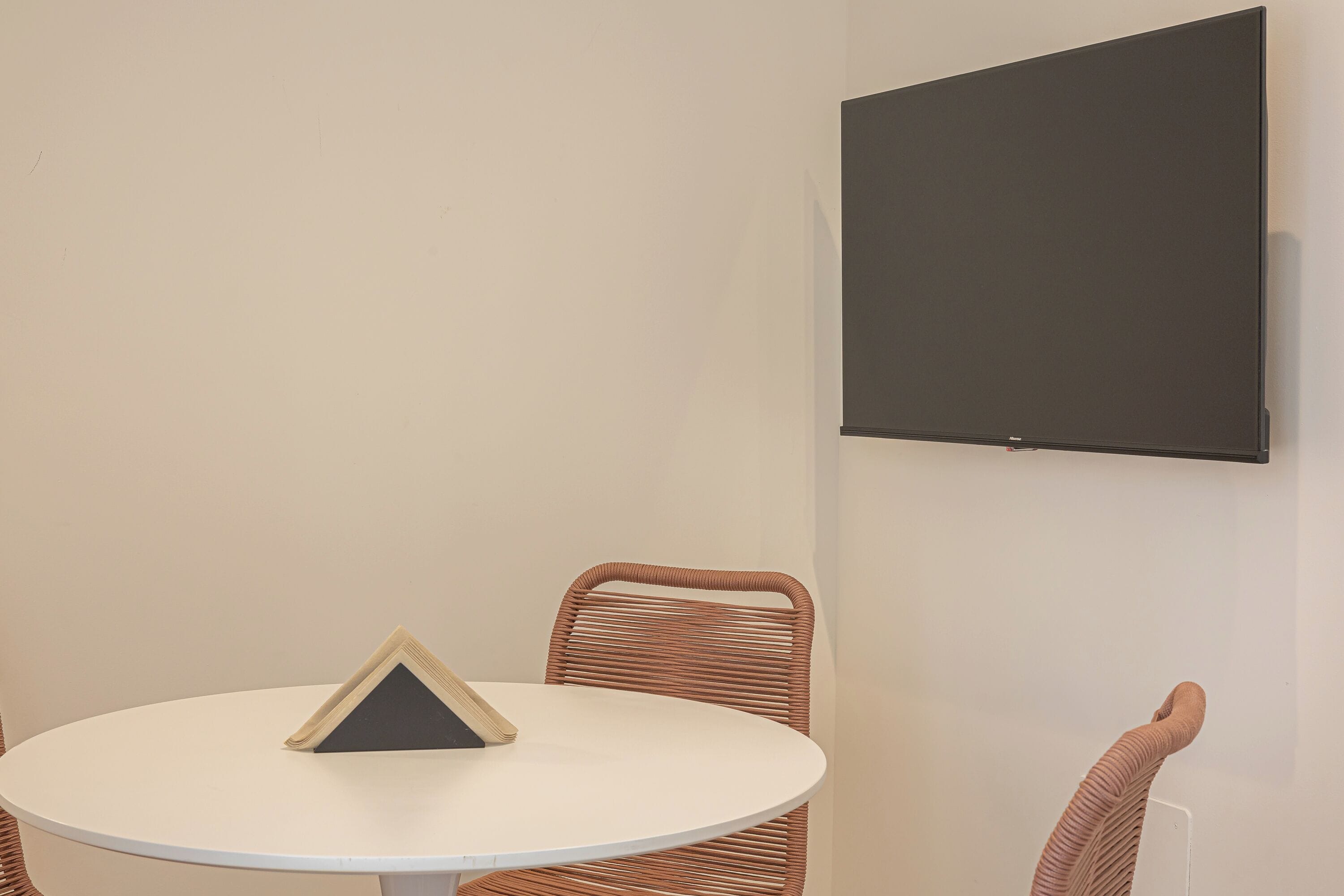 Una mesa redonda con un objeto triangular encima, una silla de mimbre y un televisor fijado a la pared.