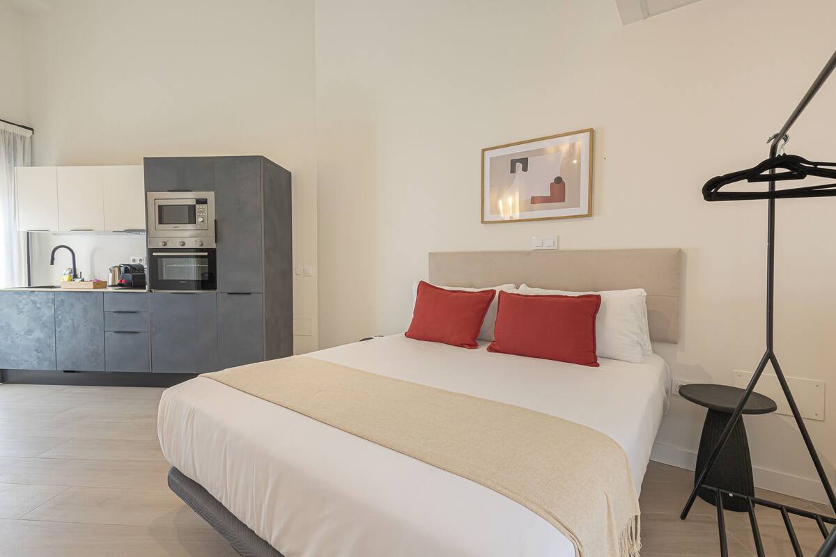 A modern hotel room with a large bed, a kitchenette with a microwave and oven, a small dining table, and a hanging coat rack.