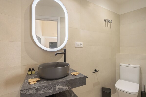 Une salle de bain avec un lavabo rond, un miroir avec éclairage intégré, des toilettes et une poubelle.