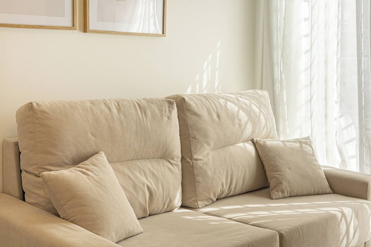 A beige sectional sofa with two cushions, positioned in a room with a plain wall and a window with sheer curtains.