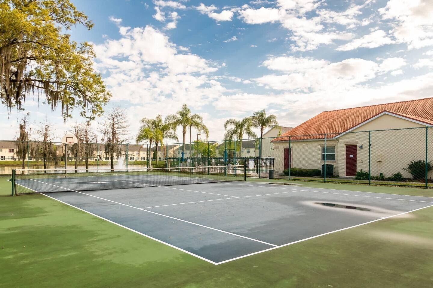 An outdoor tennis court with a net, surrounded by a fence and palm trees.