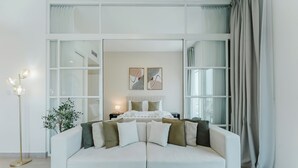 A modern living room with a white sofa, a floor lamp, and a bed with a white headboard. - PVH - 2BR Golfville Dubai Hills (Dubai)