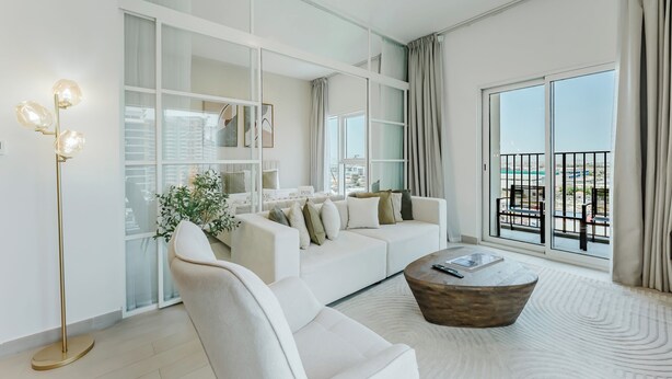 A modern living room with a white sofa, a coffee table, a floor lamp, and large windows with a view of the outdoors. - PVH - 2BR Golfville Dubai Hills (Dubai)