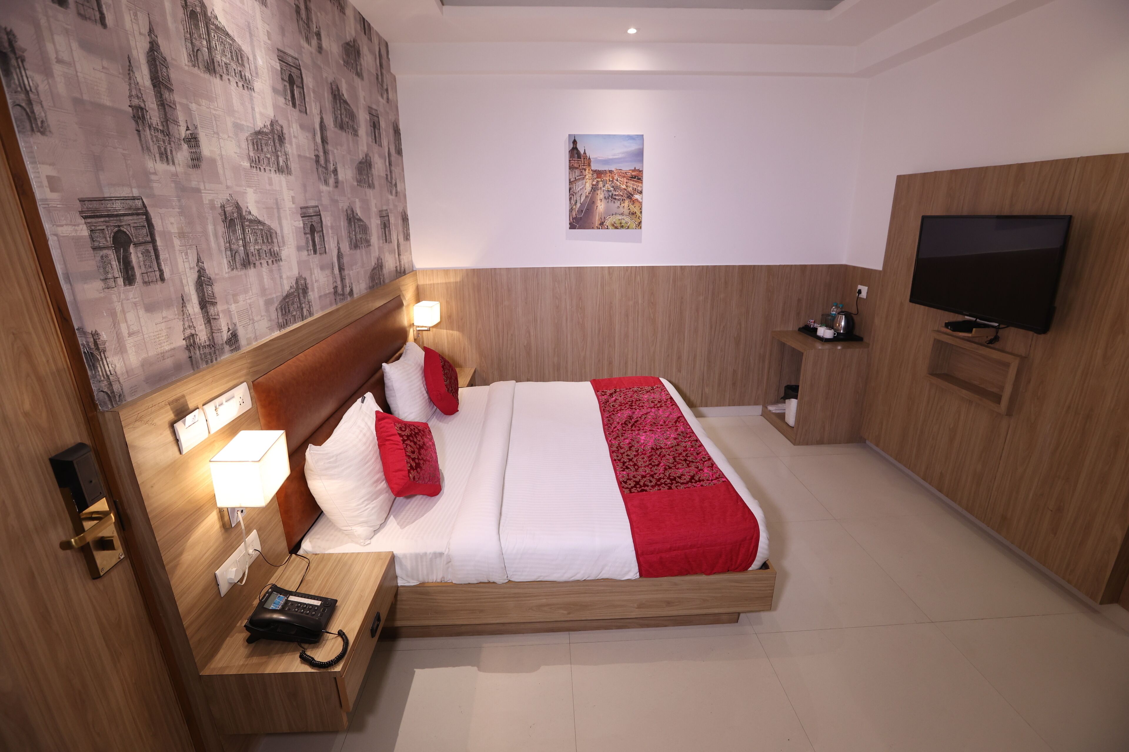 A hotel room with a wooden bed, a bedside table with a lamp, a telephone, a TV, and a wall with a cityscape picture.