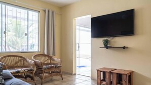 Ein Wohnzimmer mit einem großen Fenster, Korbsesseln, einem an der Wand montierten Fernseher und einem kleinen Holztisch.