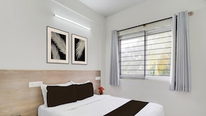 Quarto com cama, mesa de cabeceira com telefone, janela com persianas e dois desenhos de folhas emolduradas na parede.
