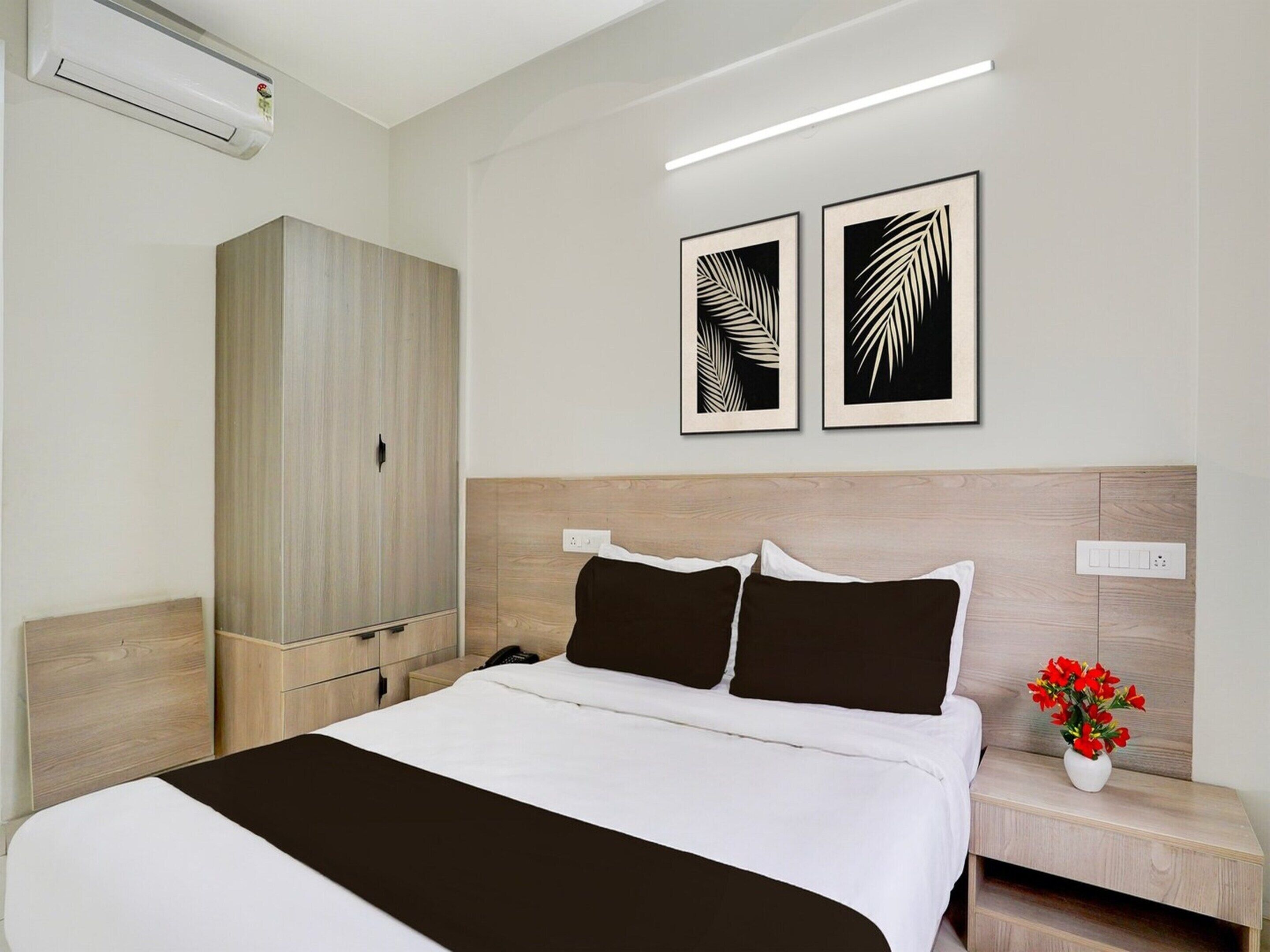 A hotel room with a bed, a wardrobe, two wall-mounted artworks, a bedside table with a vase of flowers, and a modern light fixture.