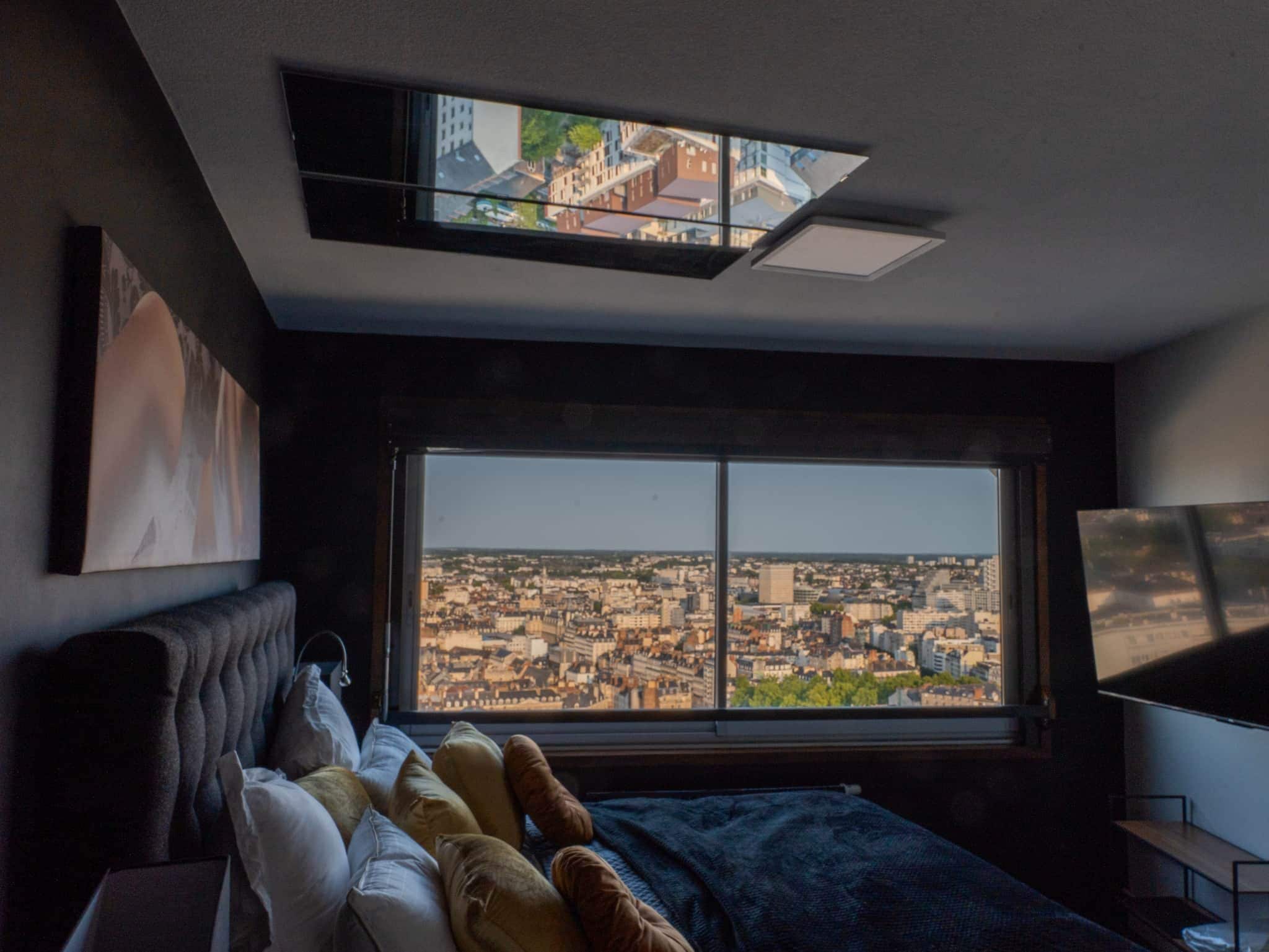 A bedroom with a large window, a bed, a TV, and a city view.