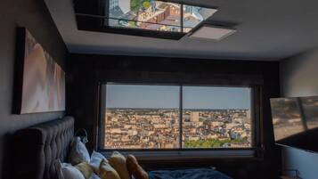 A bedroom with a large window, a bed, a TV, and a city view.