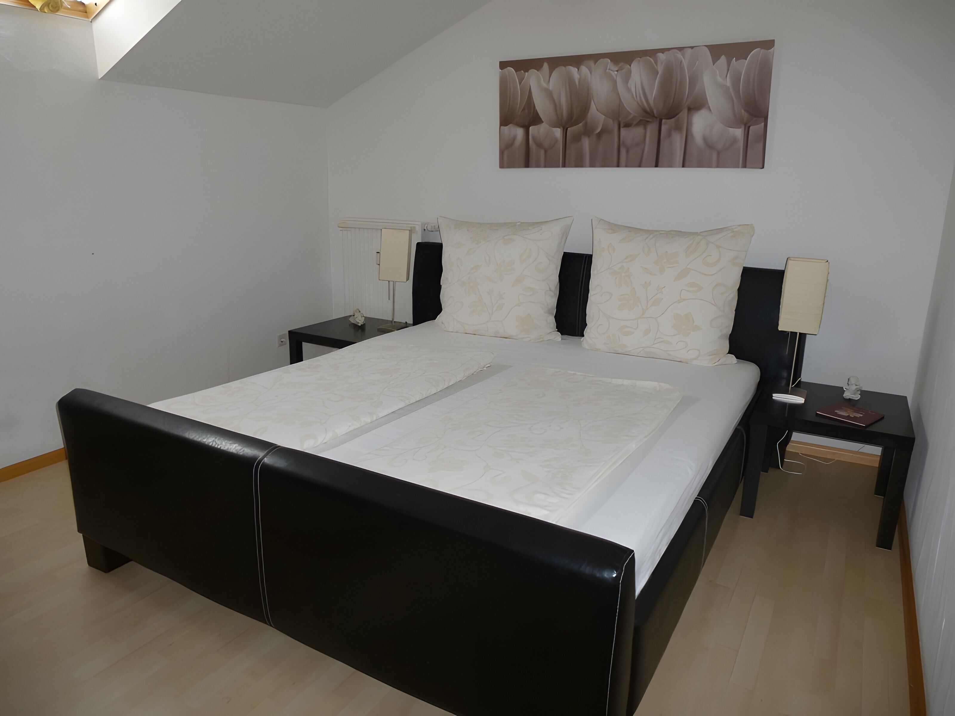 Quarto com estrutura de cama preta, roupa de cama branca, duas mesas de cabeceira com abajures, uma obra de arte na parede e teto inclinado.
