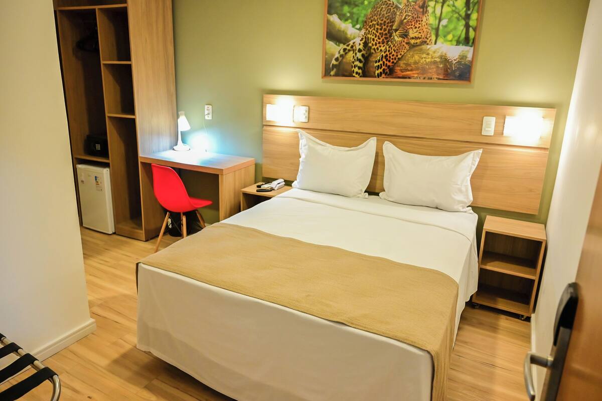 A hotel room with a bed, a desk, a chair, a wardrobe, and a painting on the wall.