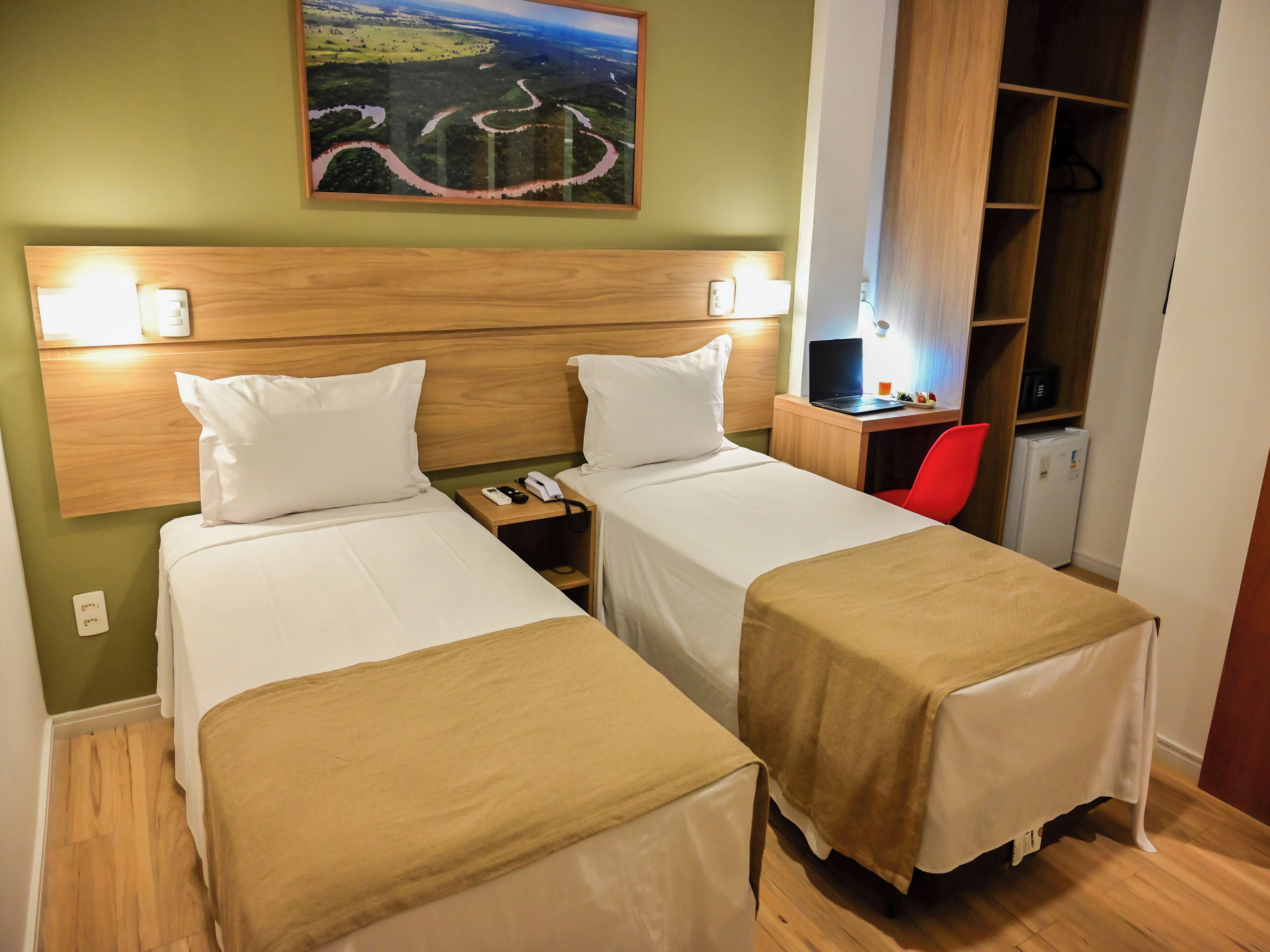 Habitación de hotel con dos camas, un escritorio con una computadora portátil, una silla roja y un cuadro en la pared.