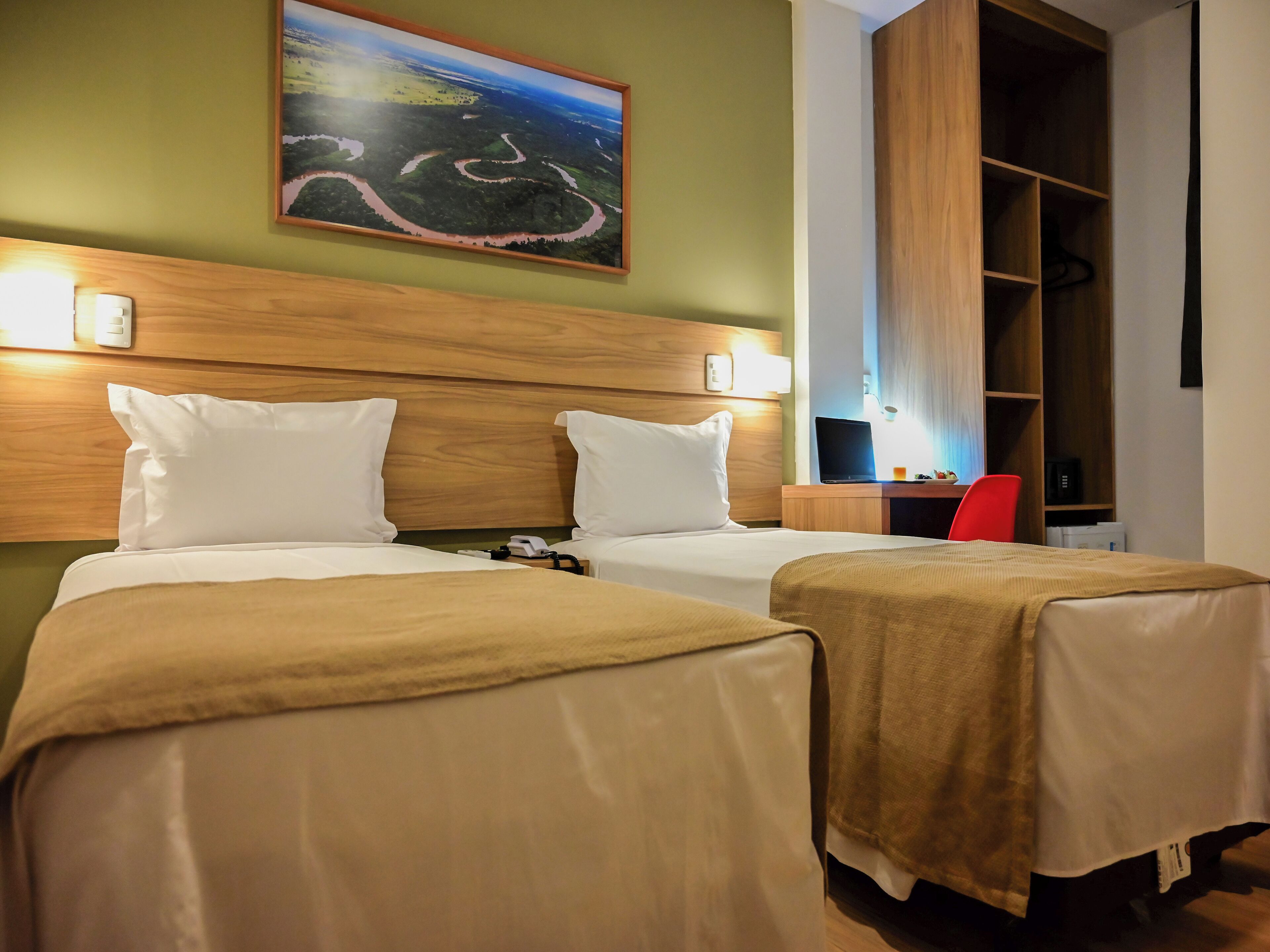 A hotel room with two beds, a desk with a laptop, a red chair, and a large painting of a landscape on the wall.