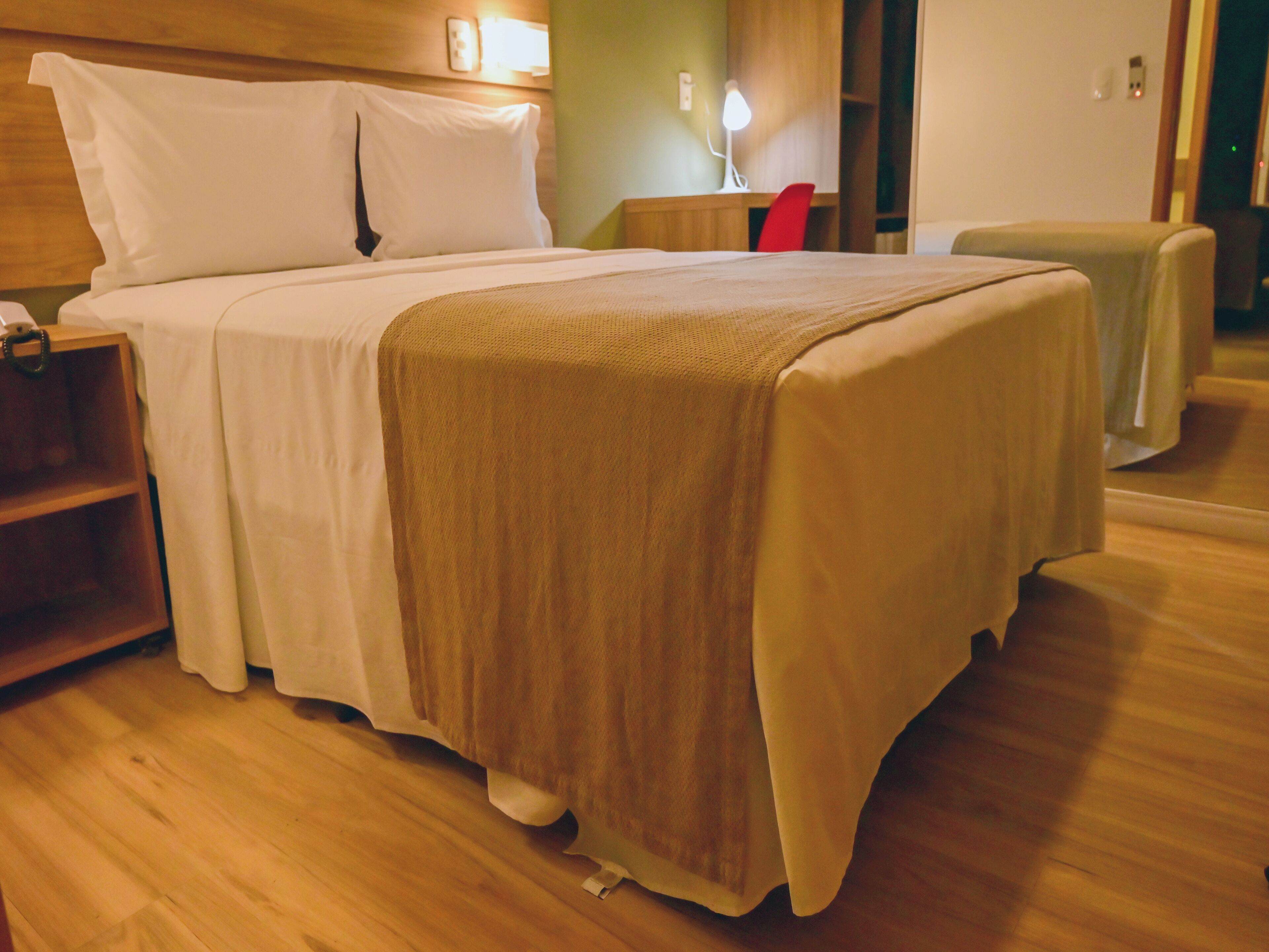 Een netjes opgemaakt bed met een wit en beige dekbedovertrek, een houten hoofdbord en een nachtkastje met een lamp.